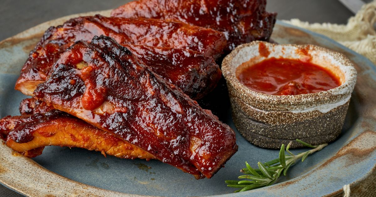 Closeup photo of a plate of cooked ribs covered with barbecue sauce with a small dish of barbecue sauce beside them