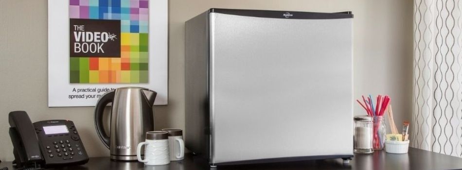 Small stainless steel refrigerator on a dark wooden office counter with a kettle and phone beside it