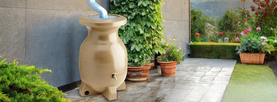 Koolscapes rain barrel set up on a stone pathway under a white downspout with a concrete wall behind and planters, grass, and a hedge around it