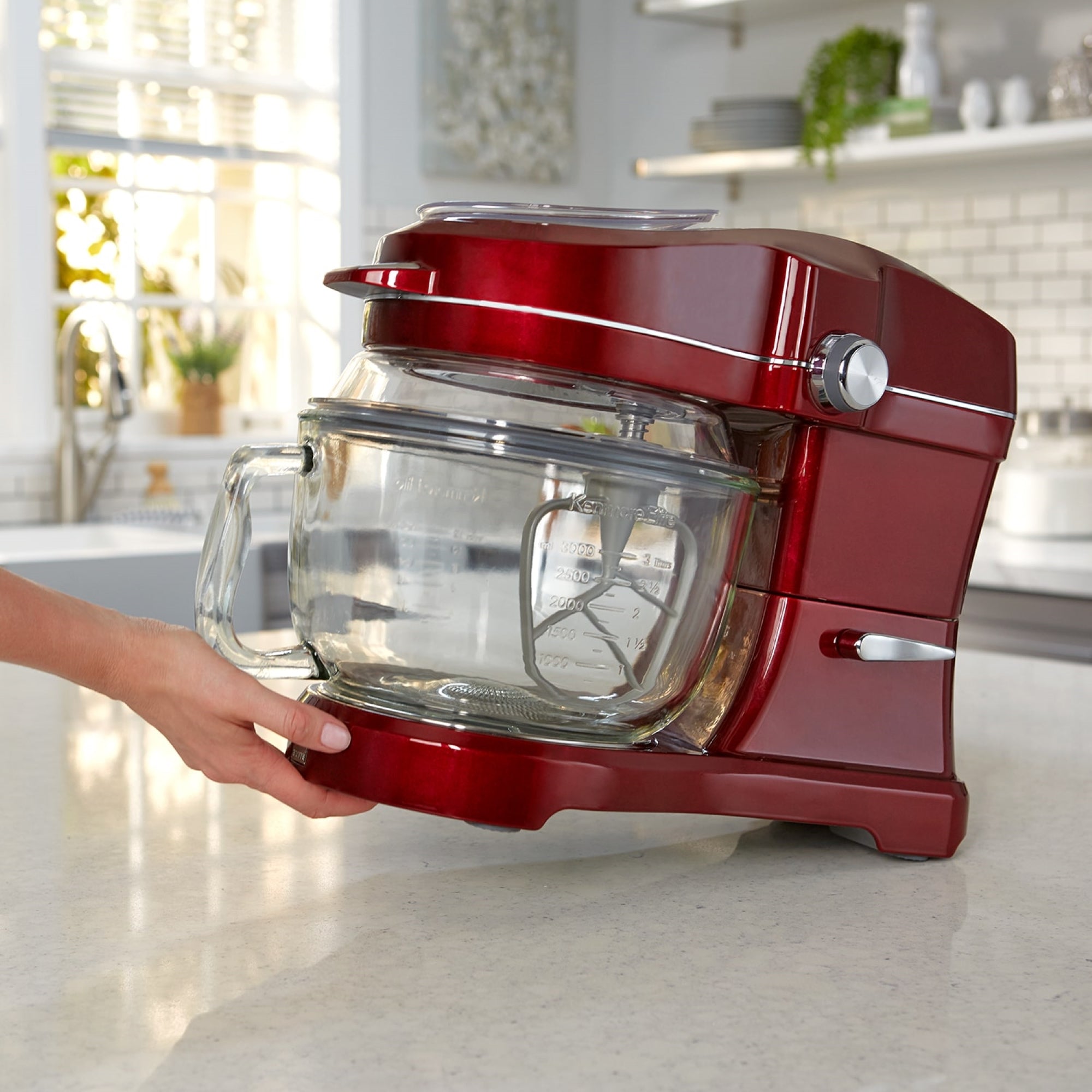 A person's hand tilting the Kenmore Elite Ovation stand mixer to move it using the tilt-and-glide feature across a light-colored kitchen counter with cupboards in the background