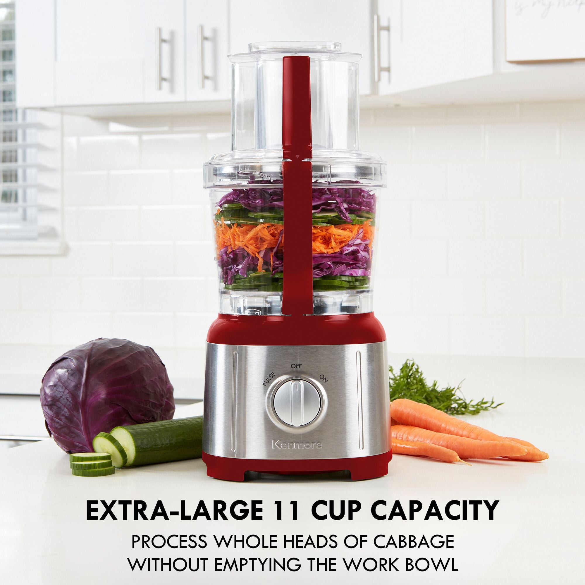 Kenmore 11 cup food processor and vegetable chopper on a light gray countertop with white tile backsplash and cupboards behind. There are chopped vegetables in the food processor and a whole red cabbage, partially sliced cucumber, carrots, and dill on either side. Text below reads, "Extra-large 11 cup capacity: Process whole heads of cabbage without emptying the work bowl"