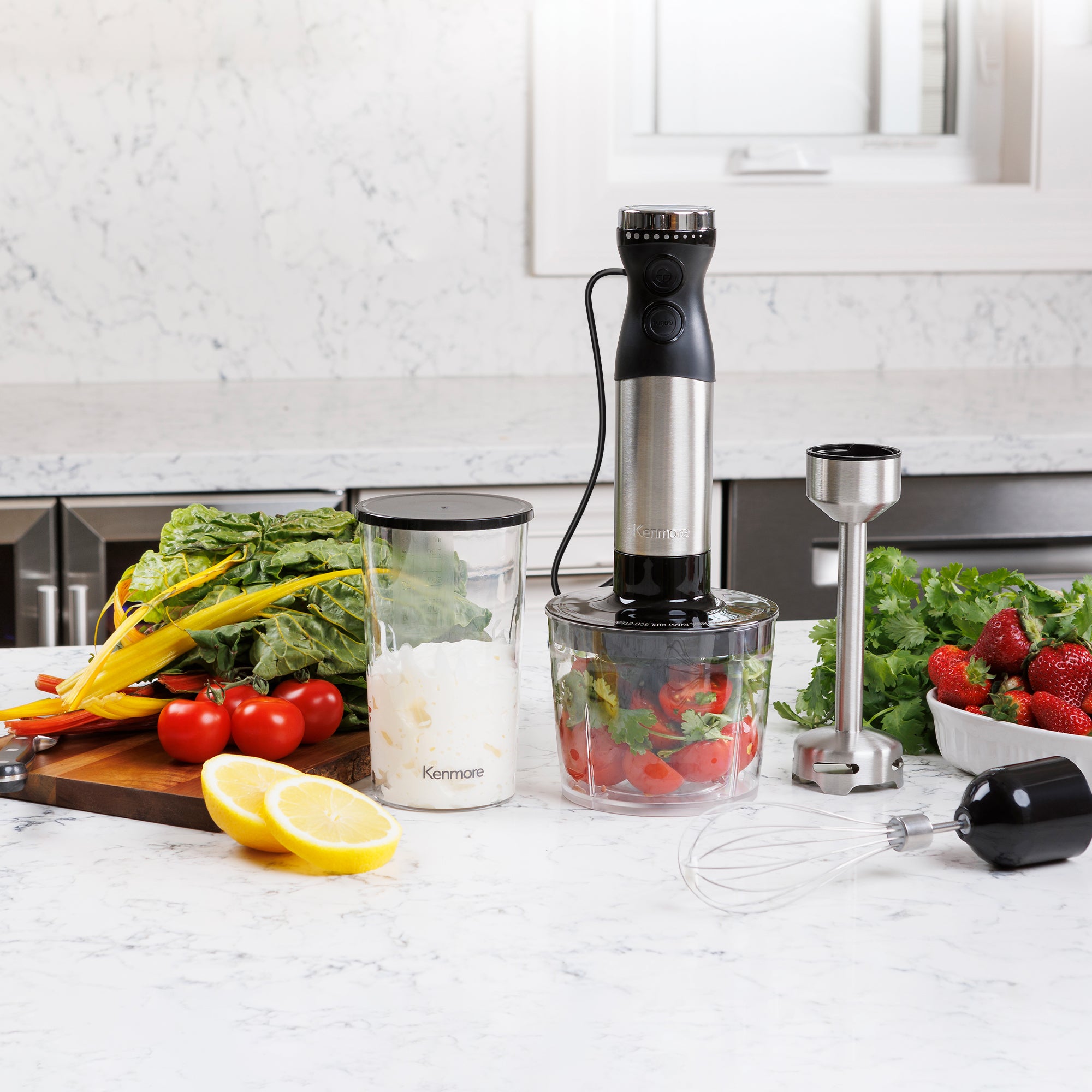 Kenmore hand blender and attachments surrounded by vegetables, greens, fruit, vegetables, and berries on a marbled white kitchen counter