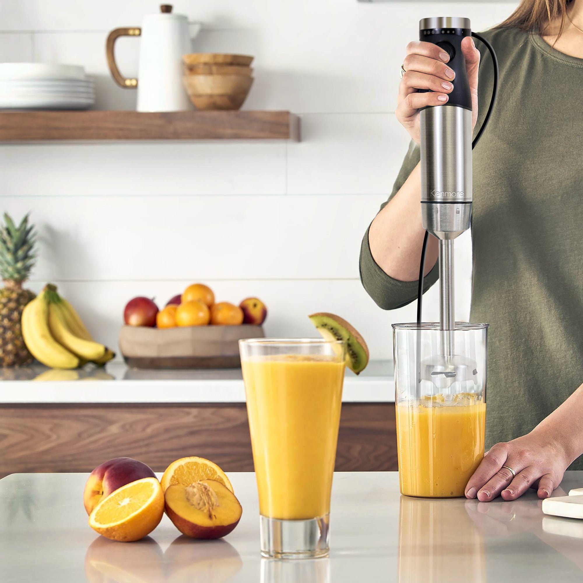 Light-skinned person in an olive-green shirt using the hand blender to puree an orange-coloured smoothie on a beige counter. There are oranges and peaches beside a glass of orange smoothie in the foreground and another kitchen counter with bowls of fruit in the background