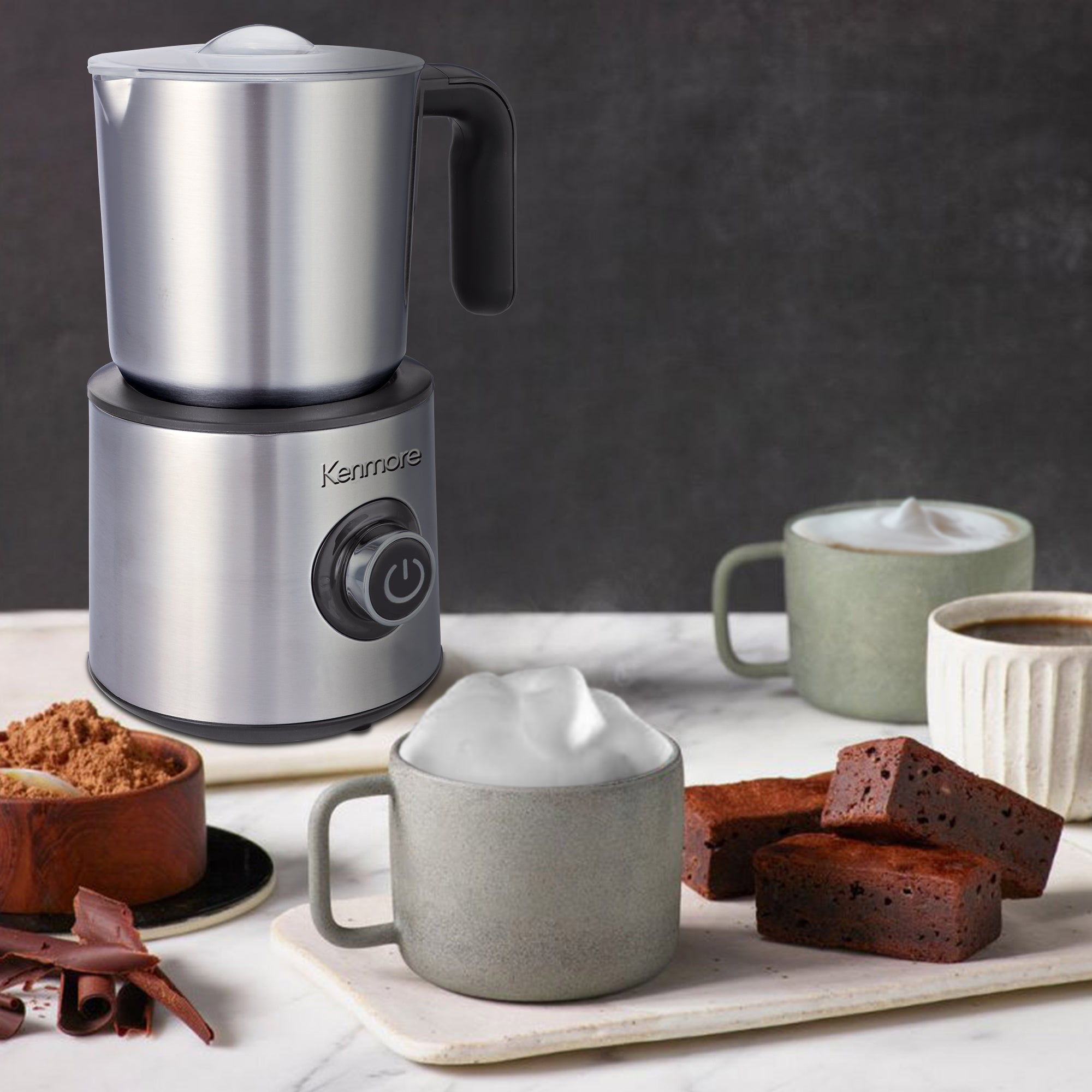 Kenmore stainless steel hot and cold foam maker on a white marble counter with a cappuccino, latte, hot chocolate, brownies, and bowl of cocoa powder arranged around it