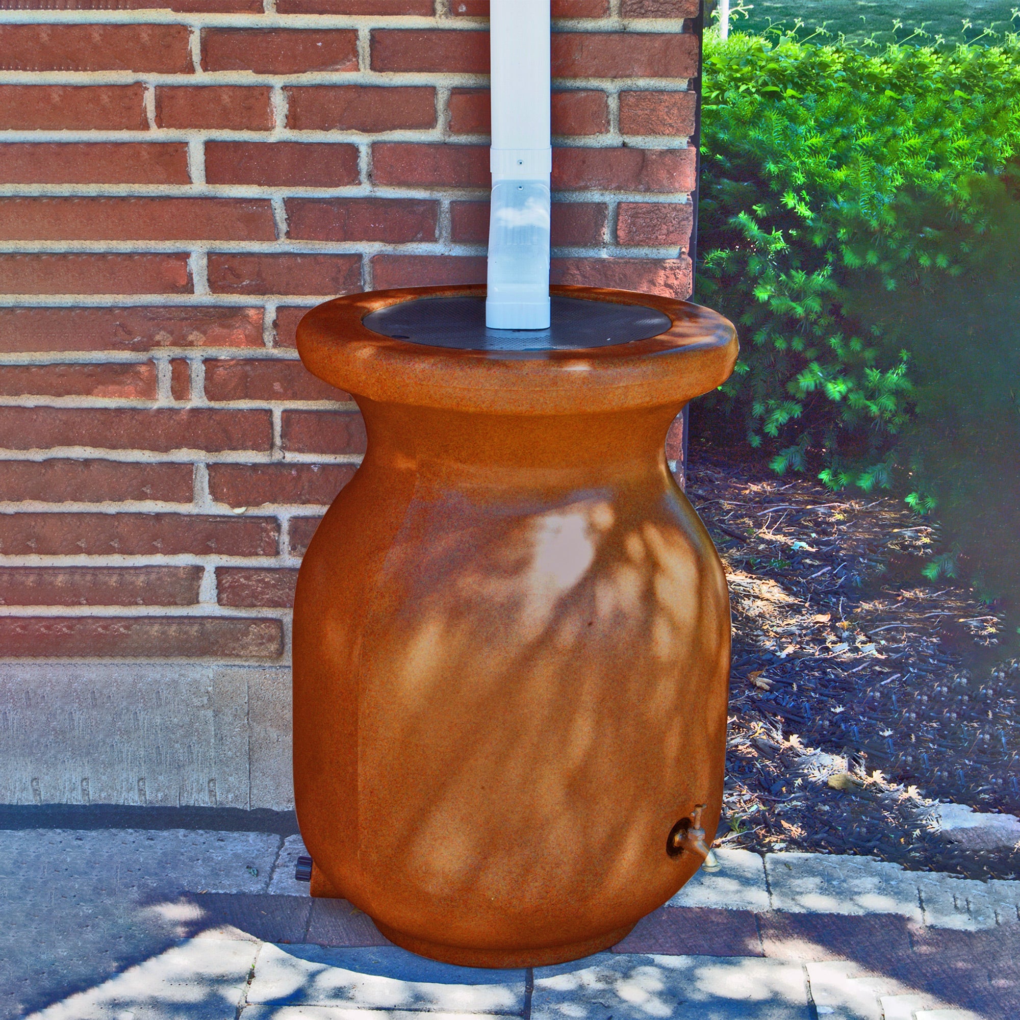 Koolscapes rain barrel set up below a white downspout with a red brick wall and a garden in the background