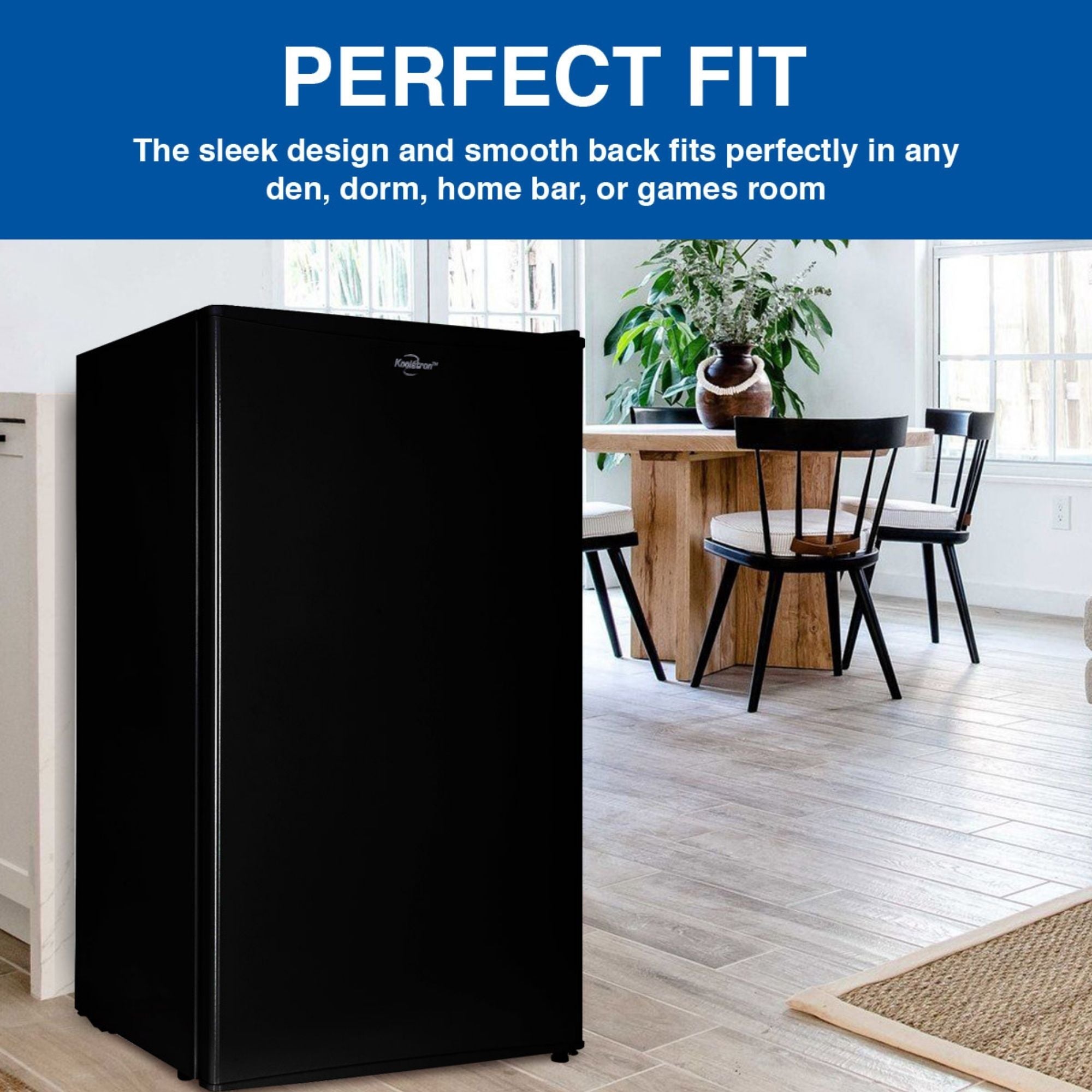 Lifestyle image of black compact fridge with freezer on a light-colored plank floor with a wooden table and chairs, two large windows, and a large potted plant in the background. Text above reads, "Perfect fit: The sleek design and smooth back fits perfectly in any den, dorm, home bar, or games room"