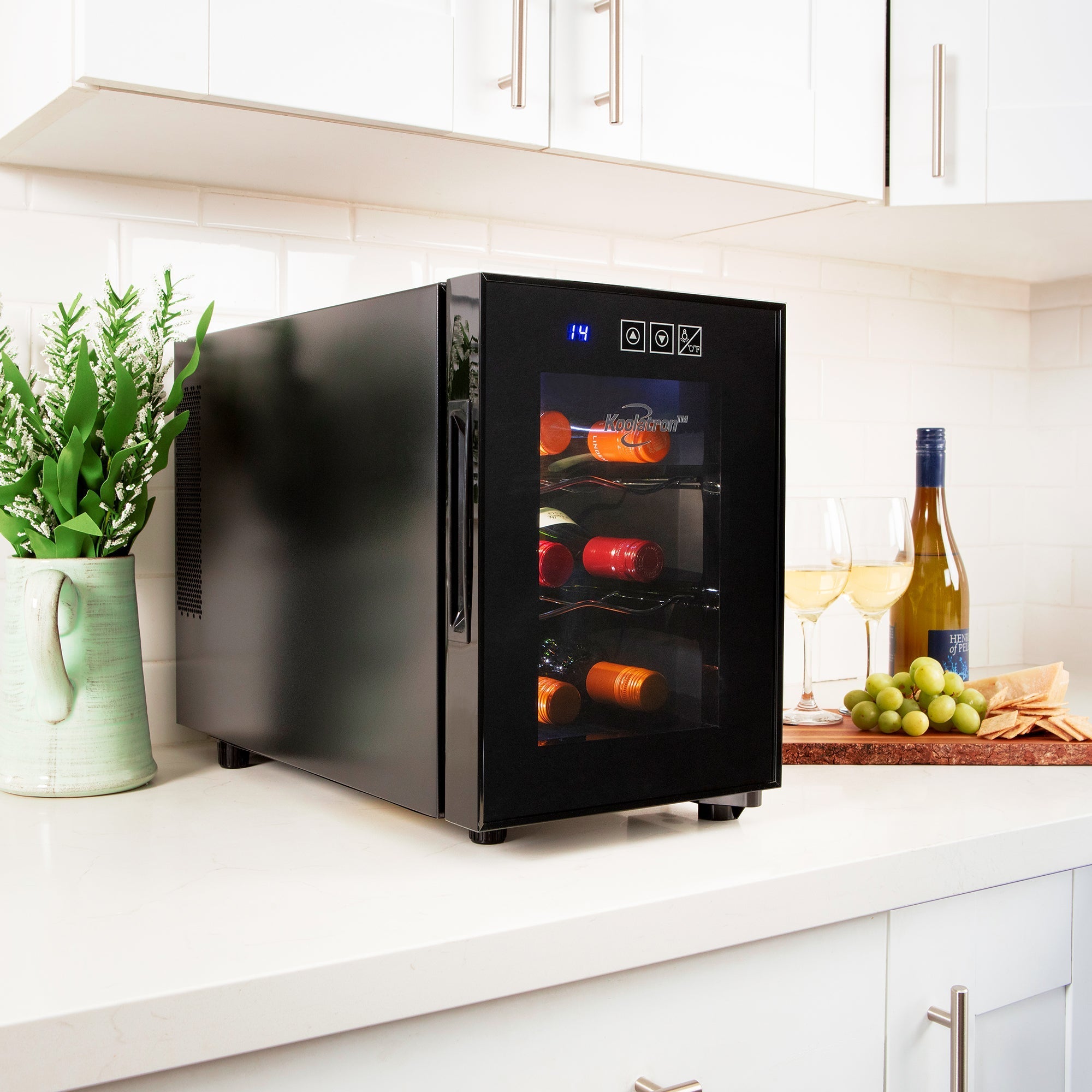 Koolatron 6 bottle wine cooler on a white kitchen counter with a light green vase of wildflowers to the left and a bottle and glasses of wine, green grapes, and crackers on a cutting board to the right