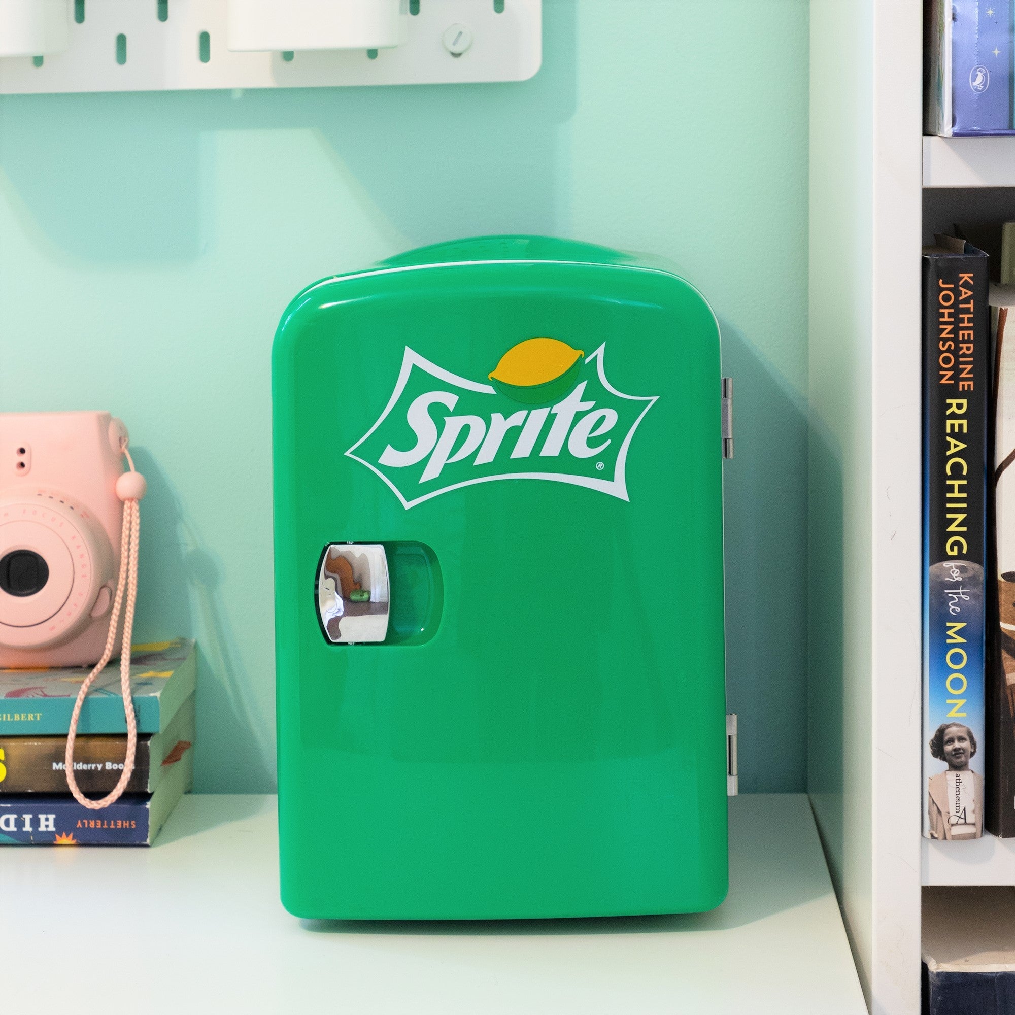 Lifestyle image of Coca-Cola Sprite 6 can mini fridge, closed, on a white desktop with an aqua wall behind. There is a stack of books and a pink camera to the left of the fridge and a bookshelf to the right
